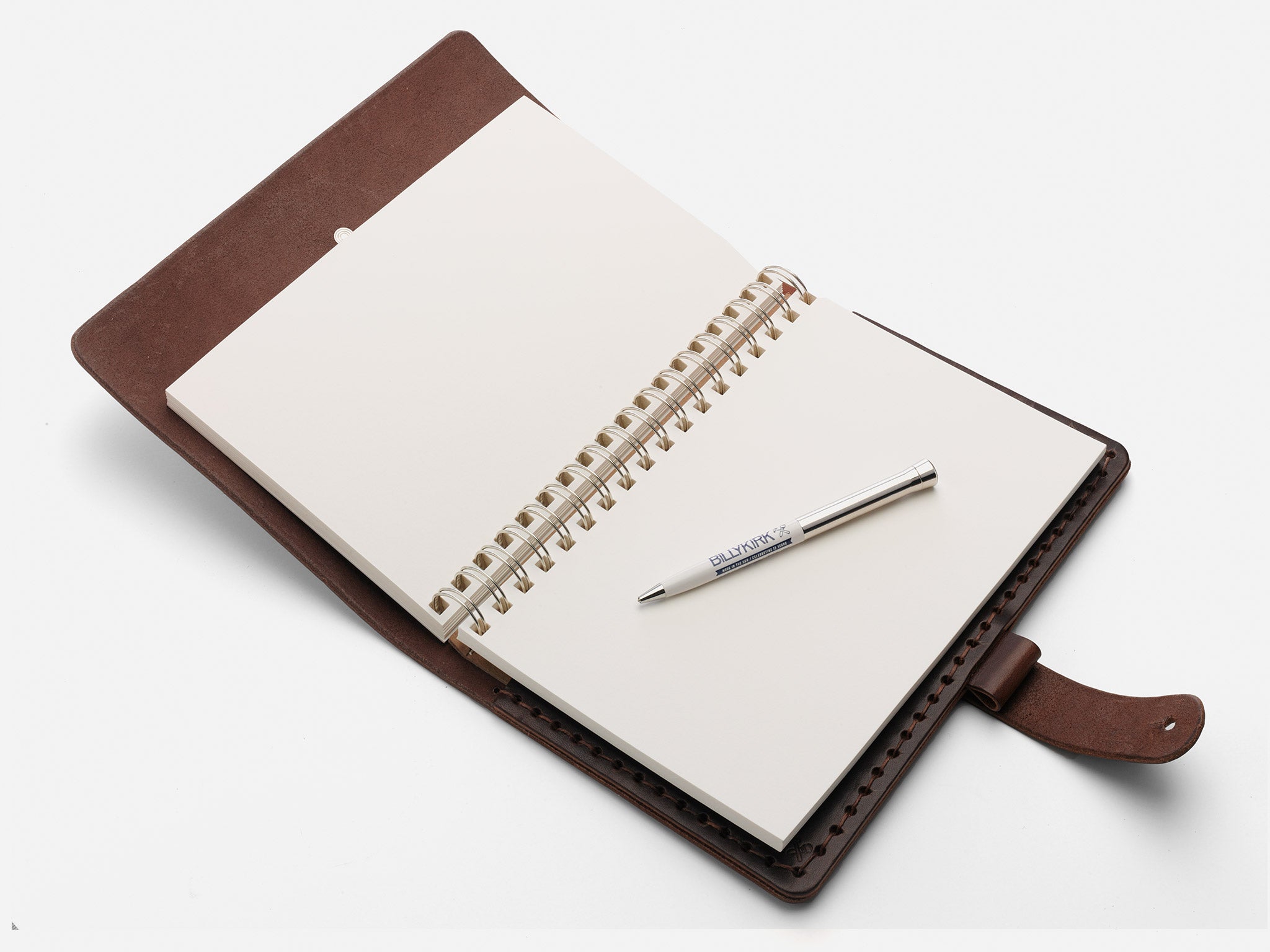 An open brown leather notebook with blank spiral pages lies on a white surface, with a silver pen placed diagonally across the pages.