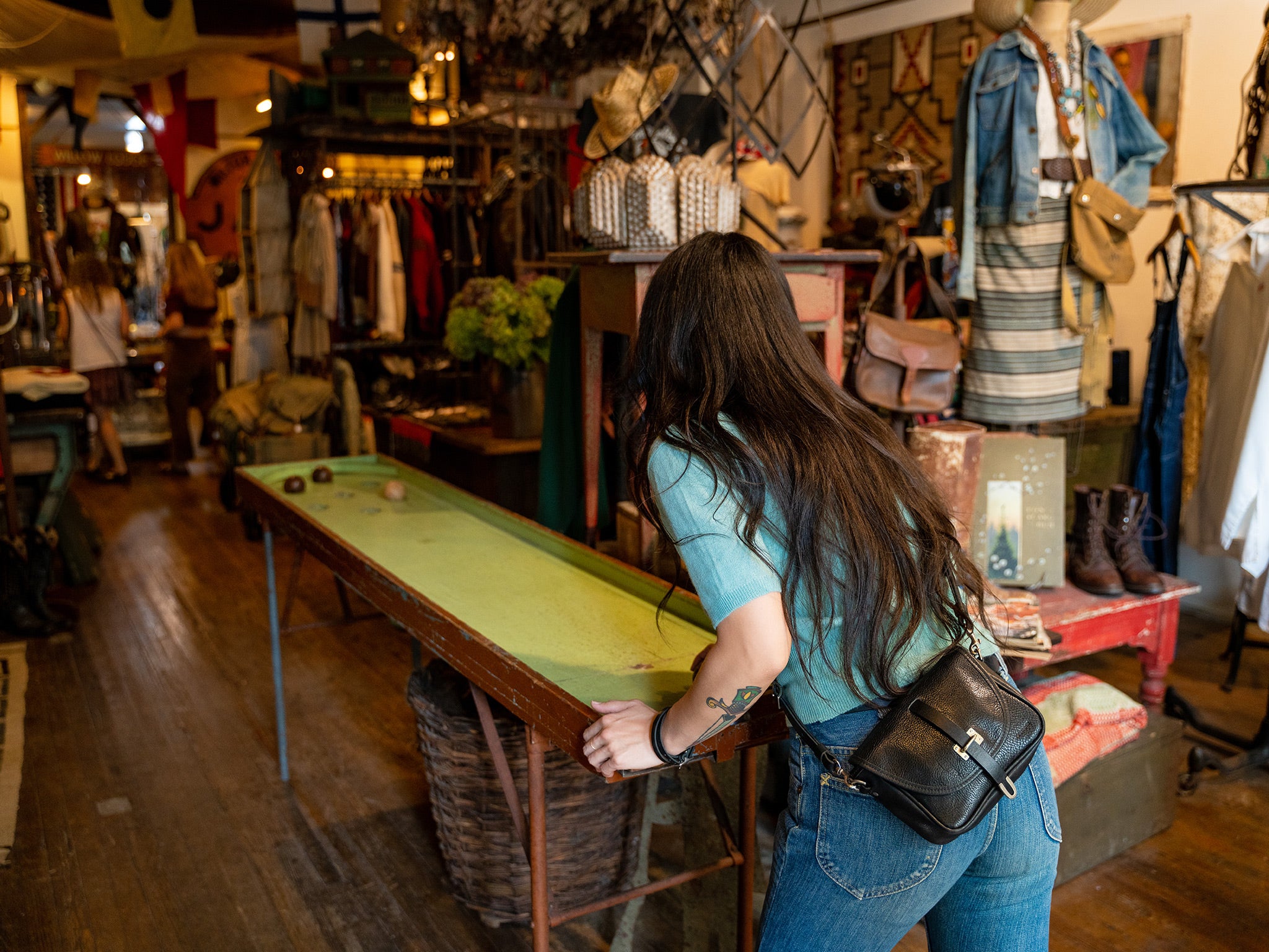 A woman with long dark hair and a black belt bag leans over a vintage shuffleboard table inside a clothing and antiques store filled with clothes, accessories, and decor items.