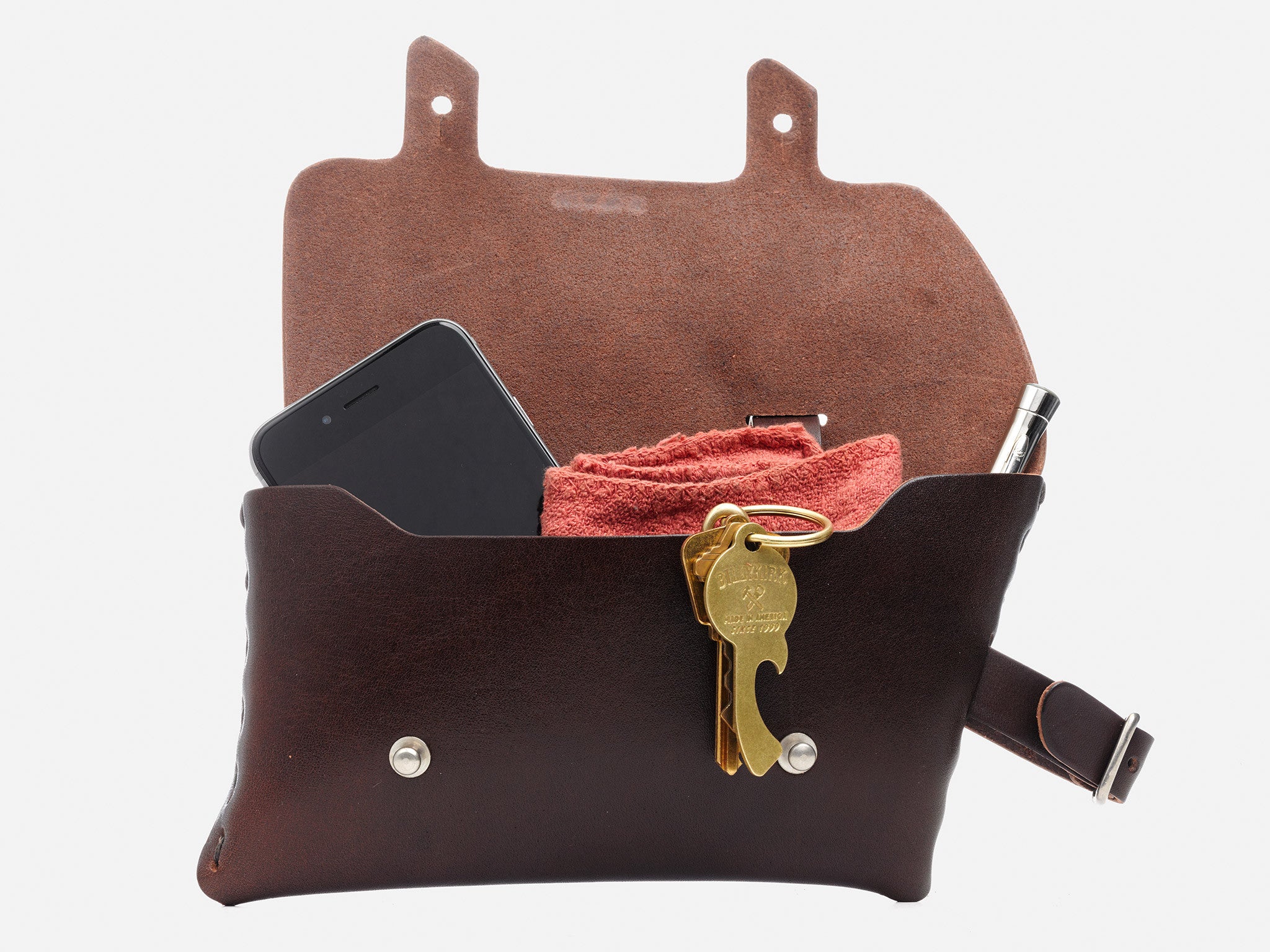 A brown leather pouch with its flap open, containing a smartphone, a red cloth, a pen, and a set of keys with a brass keychain bottle opener attached.