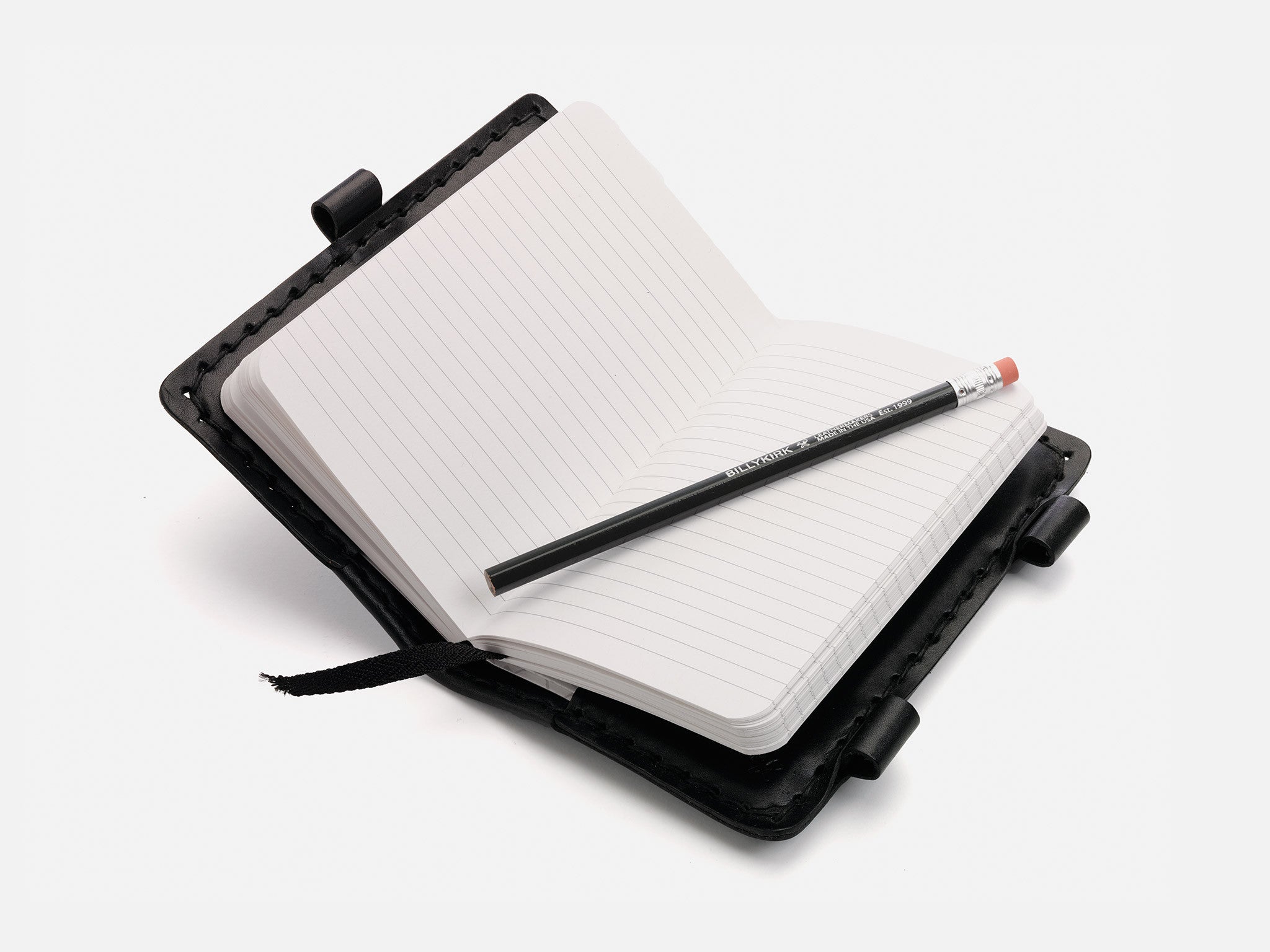 An open, lined notebook with a black leather cover lies flat, with a black pencil resting diagonally across its pages. A black ribbon bookmark is visible, and the notebook is set against a white background.