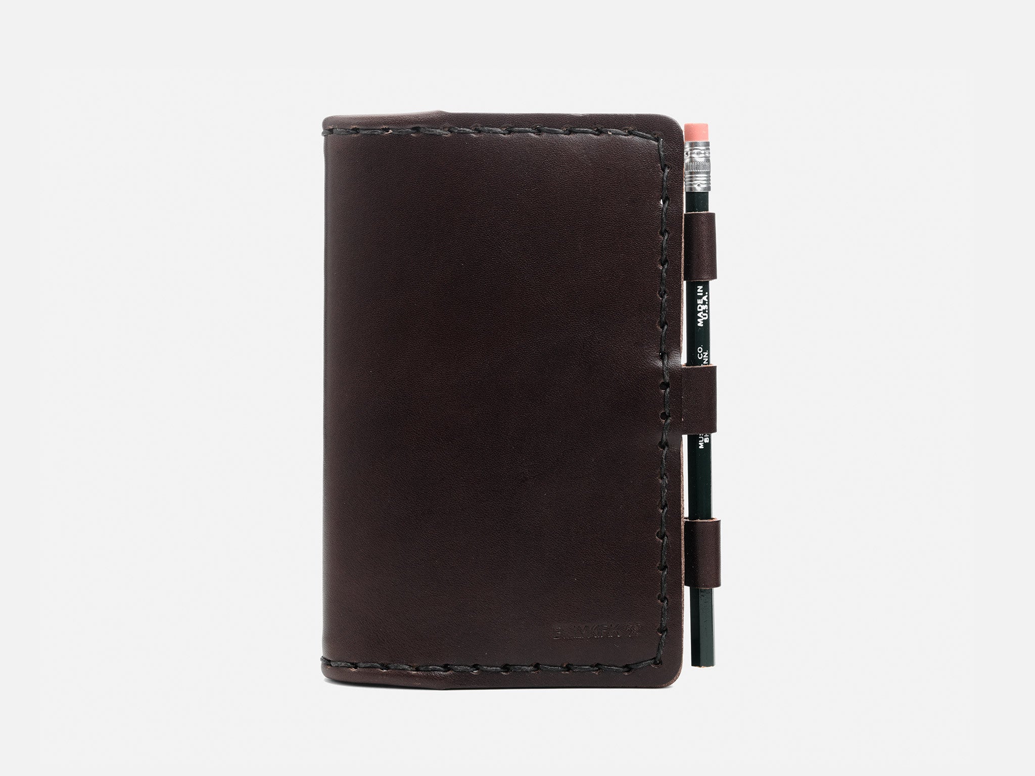 A dark brown leather notebook cover with visible stitching, closed and holding a black pencil with an eraser in an external loop on the right side, set against a plain white background.