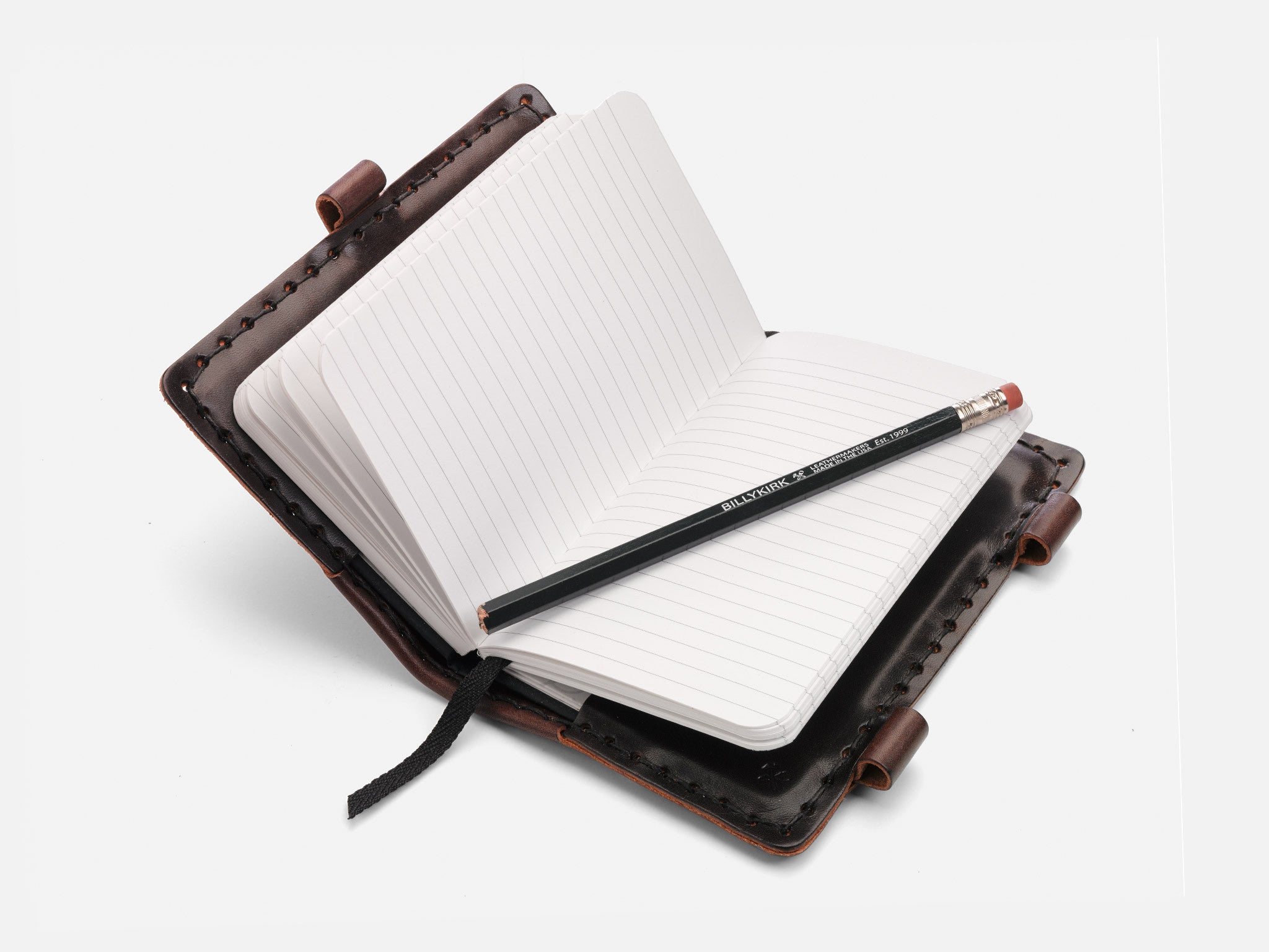 An open, lined notebook with a dark leather cover lies flat. A black pencil rests diagonally across the pages, and a black ribbon bookmark extends from the bottom.