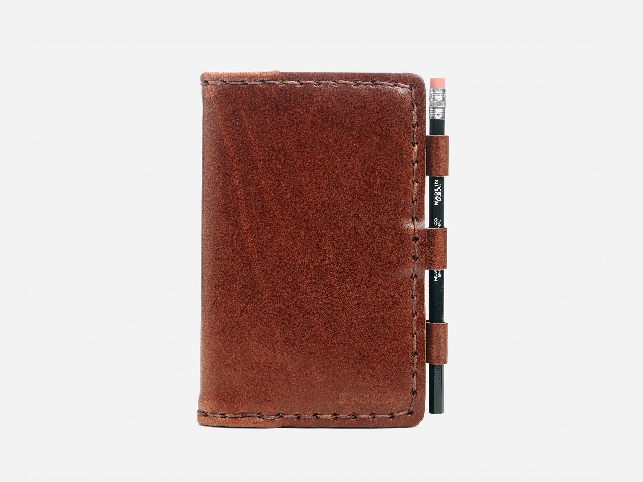 A brown leather notebook cover with visible stitching, holding a black pencil secured in three loops on the right side, set against a plain white background.