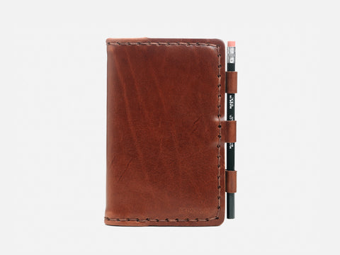 A brown leather notebook cover with visible stitching, holding a black pencil secured in three loops on the right side, set against a plain white background.
