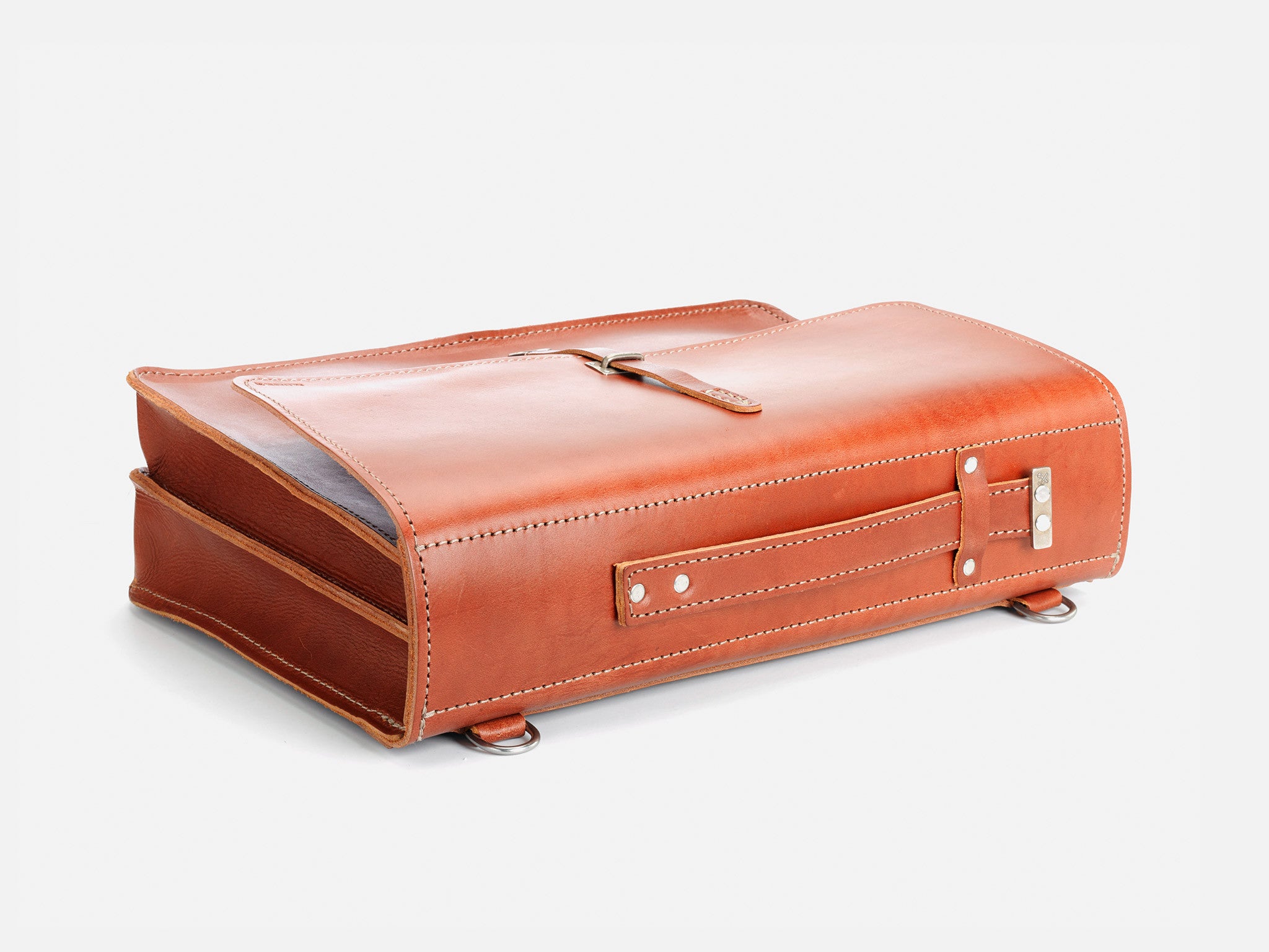 A brown leather briefcase with white stitching, metal studs on the bottom, and a strap closure, lying flat on a white background.